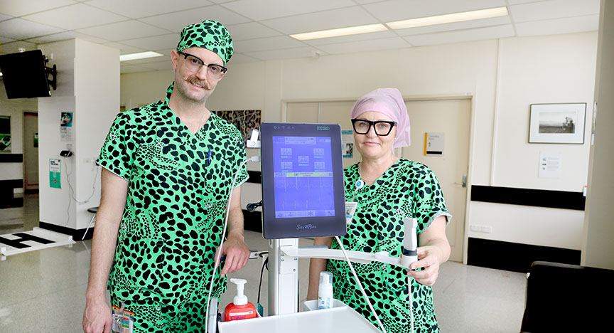 Two healthcare workers in green scrubs stand beside medical equipment in a hospital room.