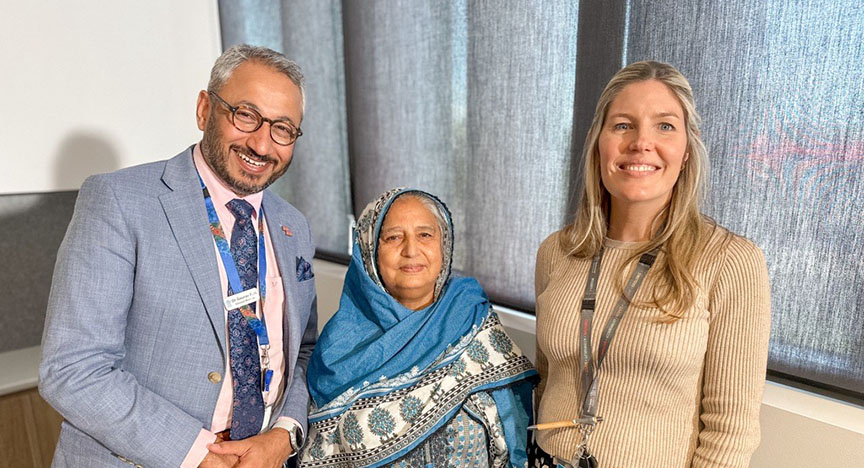 Three people stand together in front of a window with grey curtains. The person in the middle wears a blue and white patterned shawl.