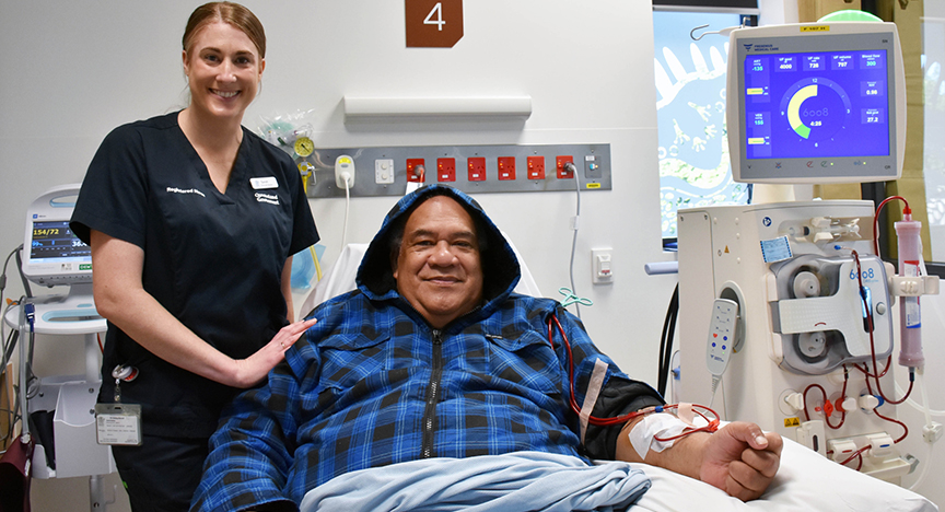 A healthcare worker stands beside a patient undergoing dialysis treatment in a hospital room.