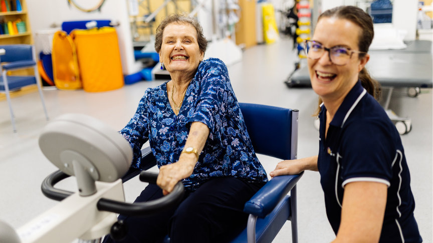 Grey haired woman sitting on a chair in focus next to younger healthcare worker who is slightly out of focus