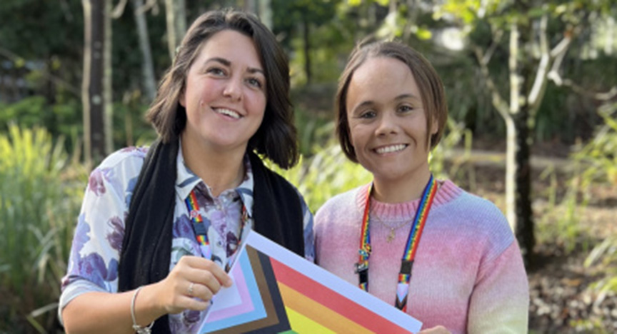 Two people holding a Progress Pride flag, wearing rainbow lanyards, standing outdoors with trees in the background.