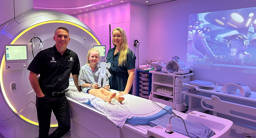 A child sits on a medical bed holding a teddy bear, accompanied by two adults in a brightly lit, futuristic medical room with animated characters projected on the wall.