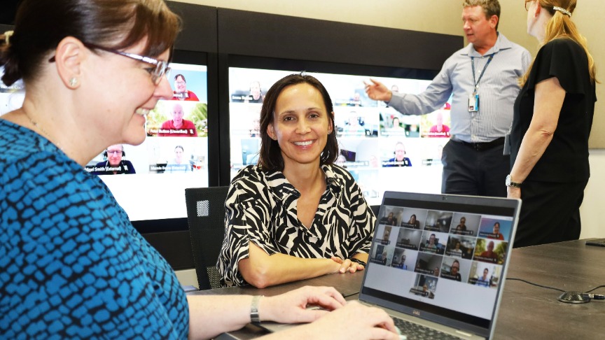 Renee Cook and a colleague sit at a table with a laptop, working on an allied health telehealth project together, while two people talk in the background