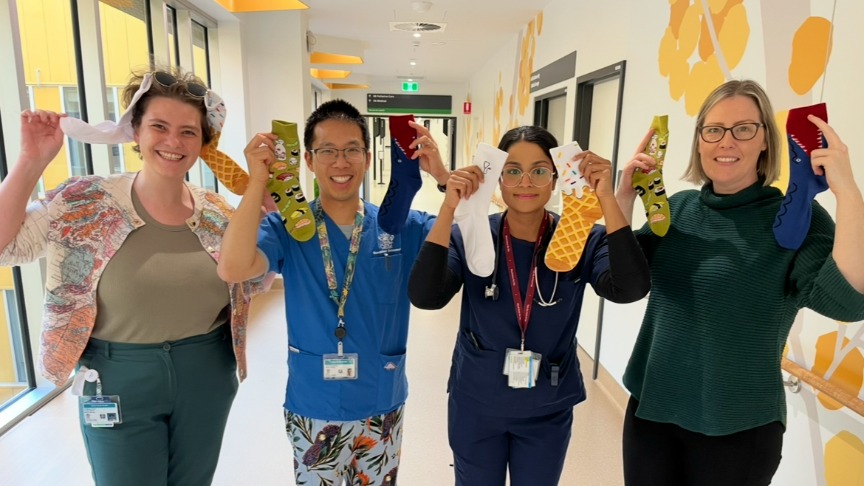 four healthcare professionals holding up crazy socs in front of a wall featuring yellow wattle