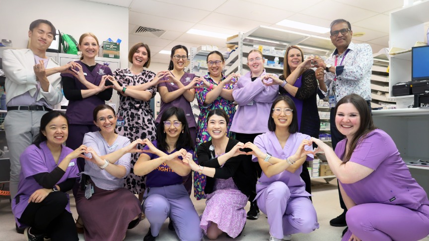 QEII Pharmacy team wearing purple and making heart hands in pharmacy.