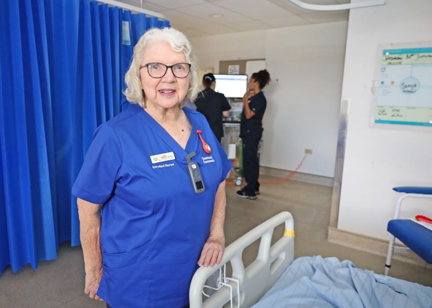 an experienced female nurse in scrubs stands near a hospital bed with other nurses in the background