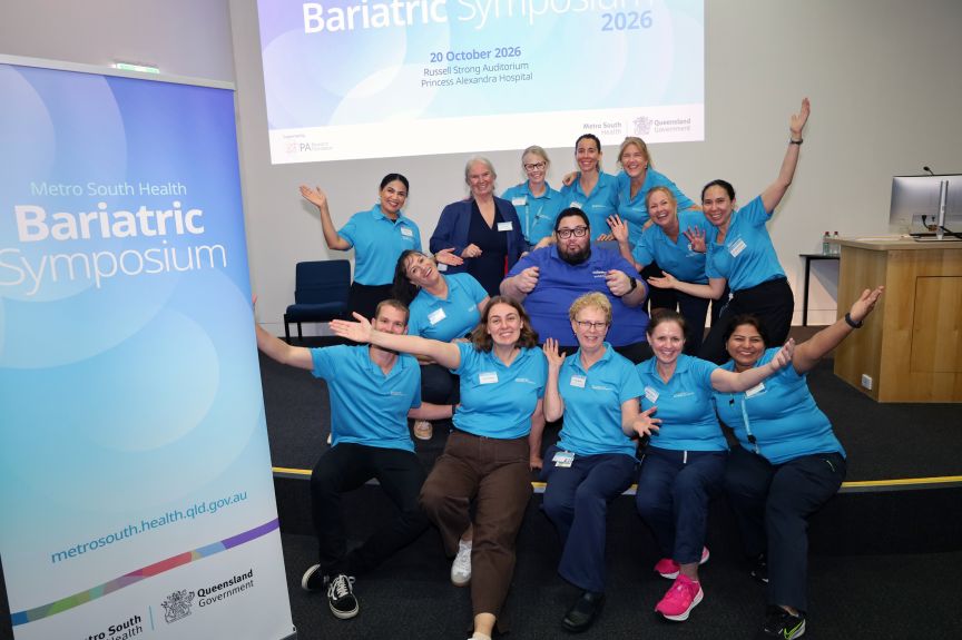 A group of men and women in blue shirts on a stage represent the program committee for a clinical symposium