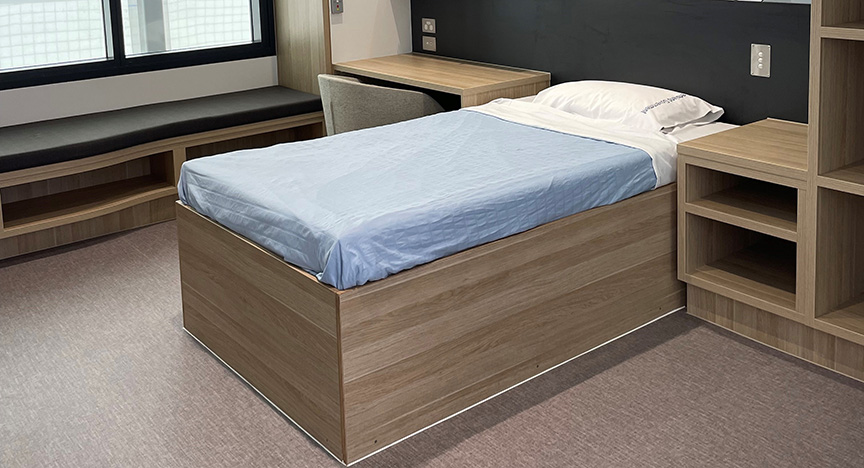 A single bed with a light blue sheet in a modern, minimalistic room with a desk, chair, and shelving unit.