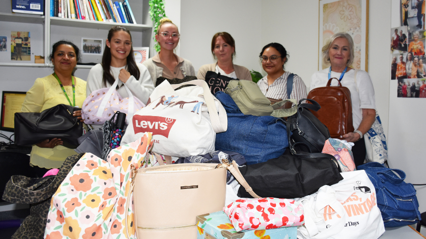 A large pile of bags in focus in the foreground on a table, with a group of five women standing behind, out of focus