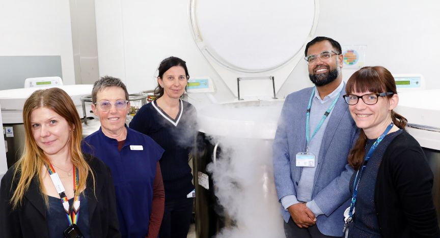 A group of people stands around a cryogenic storage tank emitting vapor in a laboratory setting.