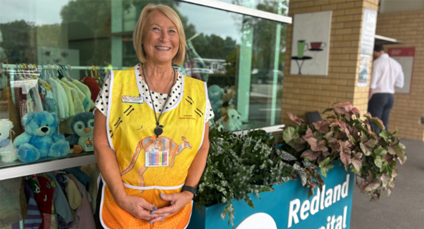 An older woman standing in front of a window at Redland Hospital wearing a bright yellow volunteer vest with a big smile on her face.