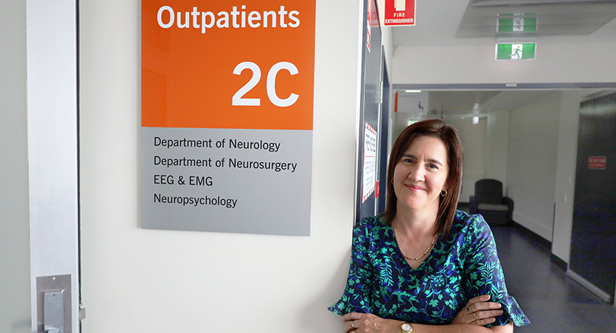 A person stands next to a sign for Outpatients 2C, which includes the Department of Neurology, Department of Neurosurgery, EEG & EMG, and Neuropsychology.