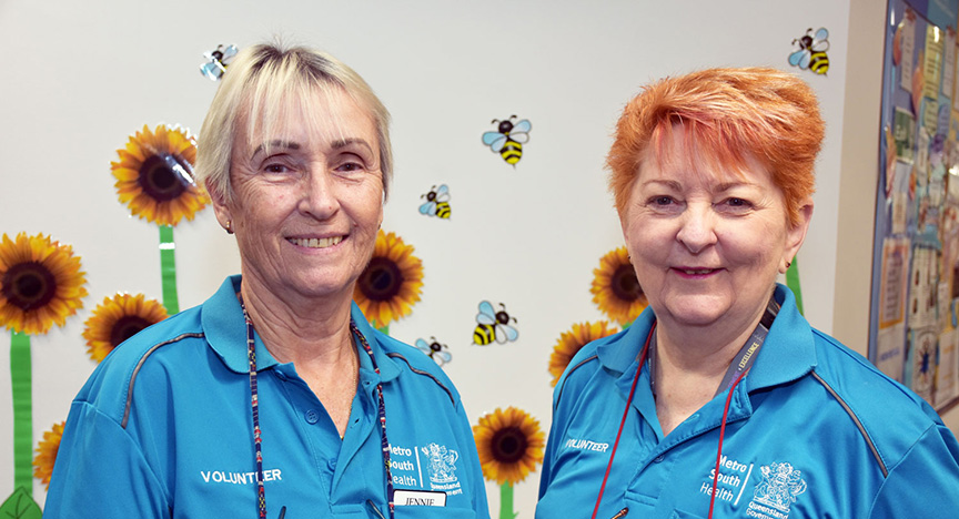 Two volunteers in blue uniforms stand in front of a wall decorated with sunflowers and bees.