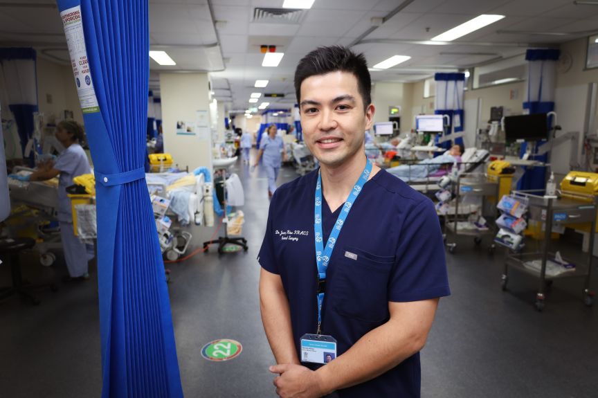A male surgeon in scrubs standing in a theatre recovery area flanked by beds, patients and nurses