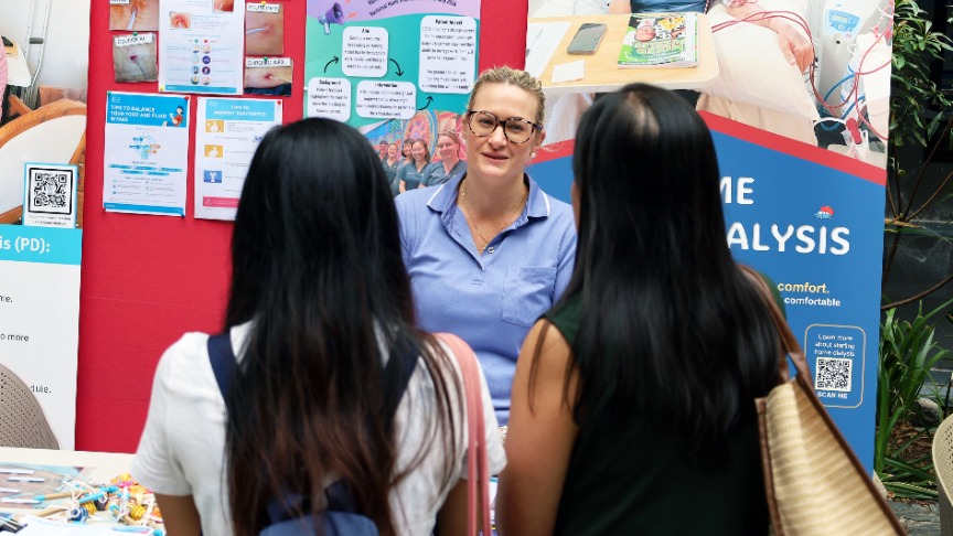 A female nurse converses with two attendees at a recruitment open day, sharing insights about nursing careers