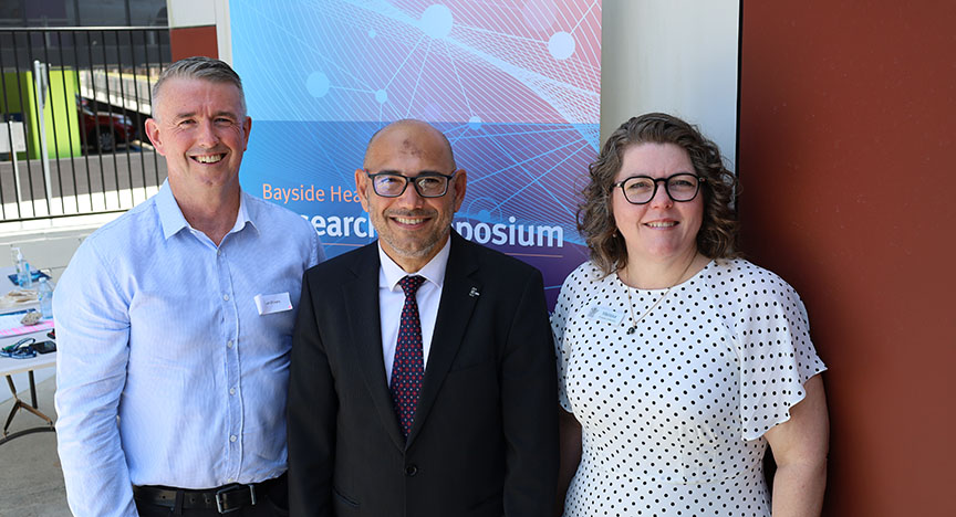 Three people standing in front of a banner that reads Bayside Health Research Symposium. The person in the middle is wearing a suit.