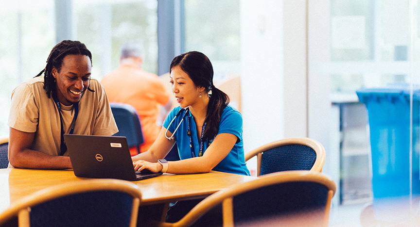 A healthcare worker and a person are seated at a table, discussing something on a laptop in a bright, modern room.