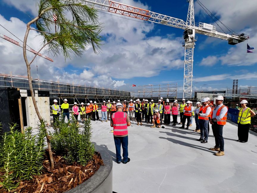A group of hospital and construction company representatives wearing high visibility protective equipment standing on the top slab of a large construction with cranes in the background and a potted plant in the foreground