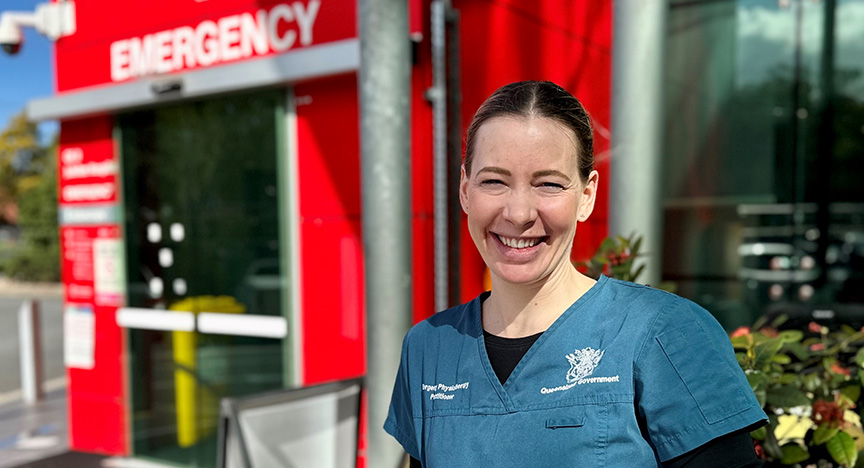 A healthcare worker in blue scrubs stands outside an emergency department entrance.