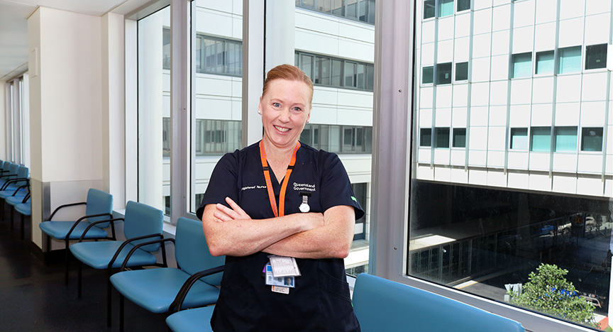 A nurse stands with arms crossed in a hospital corridor with large windows and blue chairs.