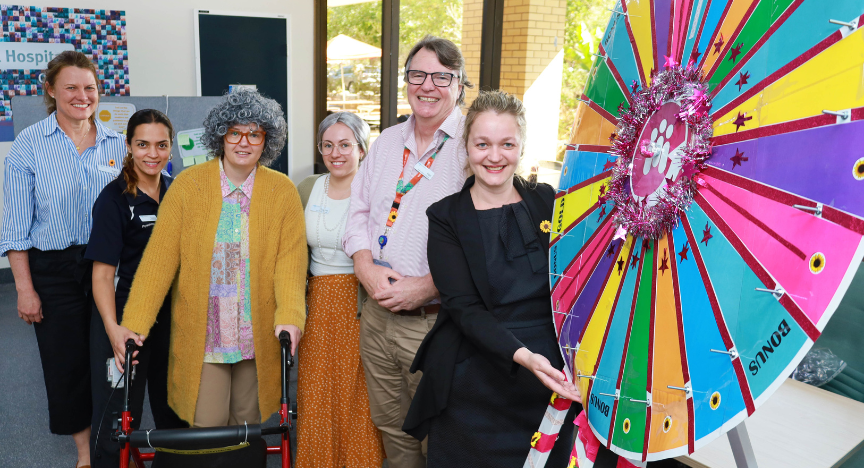 A group of people stand next to a colorful prize wheel at a hospital event. One person uses a walker.