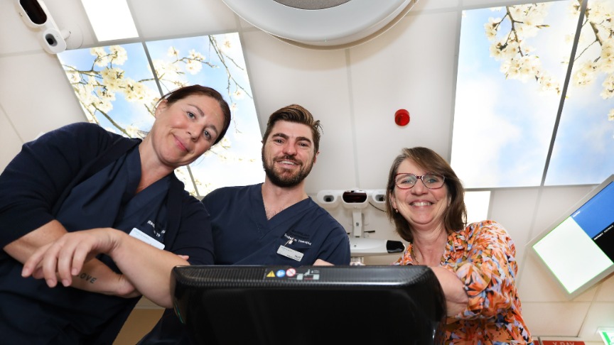 A female and a male radiation therapist alongside a female director leaning over the end of a bed platform in the renovated linear accelerator space showcasing the new illuminated ceiling tiles printed with cherry blossoms and blue sky 