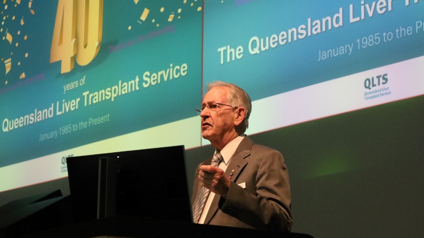 A stately man presents at a lectern in front of a large screen displaying the signage 40 Years of the Queensland Liver Transplant Service