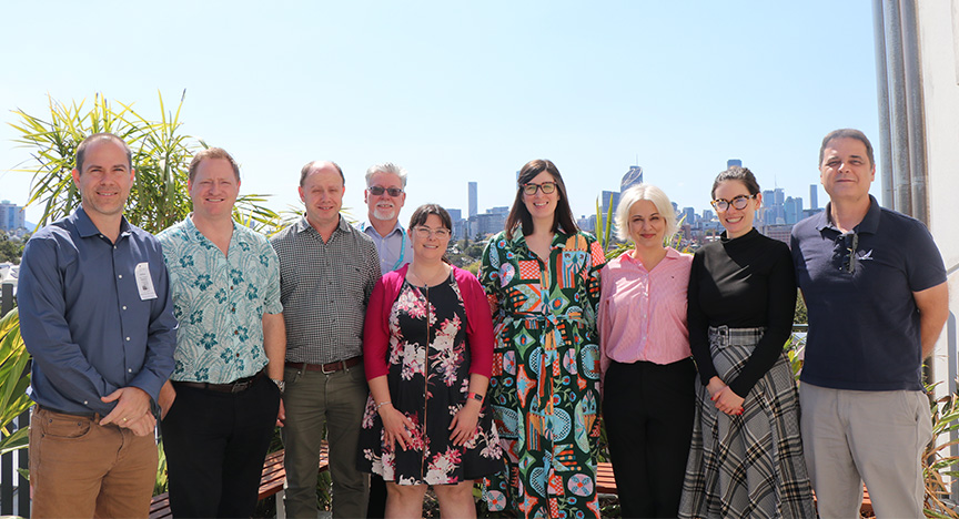 A group of people standing together smiling outside on a balcony with the blue sky and Brisbane city skyscrapers in the background.