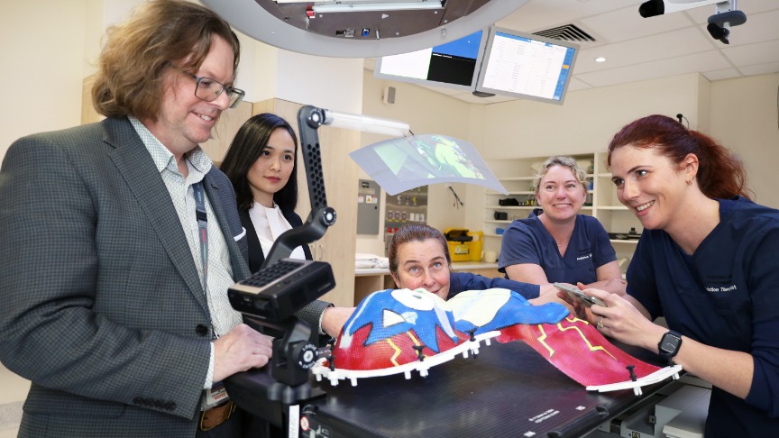 a team of five clinicians including a male doctor and four female radiation therapists and physicists set up a viewing device near a radiation therapy machine