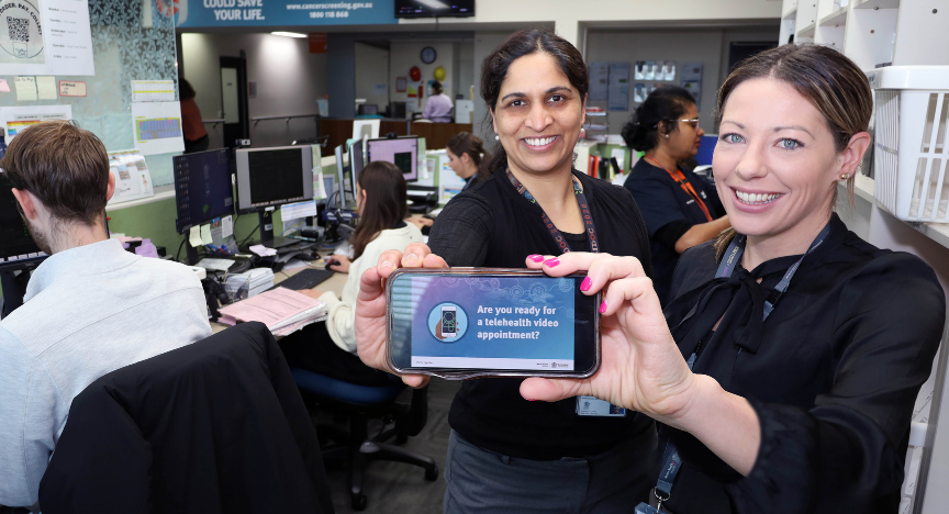 Two people in an office hold a phone displaying Are you ready for a telehealth video appointment? in front of a busy workspace with multiple computers.