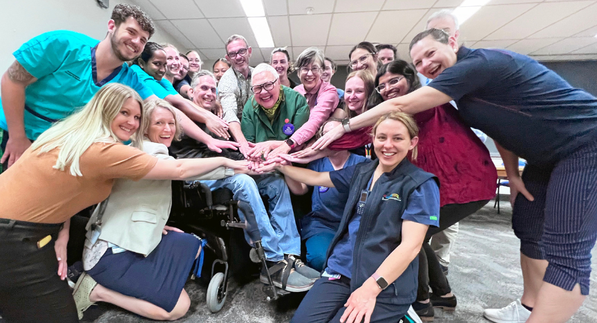 A group of people, including healthcare workers and individuals in wheelchairs, join hands in a circle in a well-lit room.