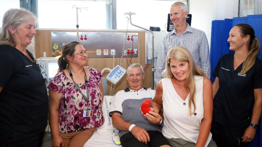 Two female nurses, a female surgeon and a male surgeon from the transplant team at PA Hospital gather around the 1000th living donor kidney transplant recipient who is a man lying in bed holding a red model of a kidney, and his living donor wife sits beside him.