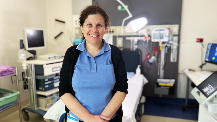 Female nurse standing in front of patient bed smiling.