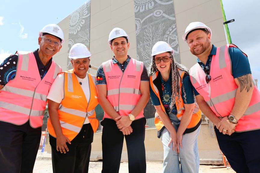 A group of Aboriginal and Torres Strait Islander men and women wearing high visibility vests and hard hats in front of a building that has artwork cladding