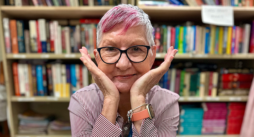 A person with short, pink-tinted hair sits in front of a bookshelf, holding their hands up to their face.