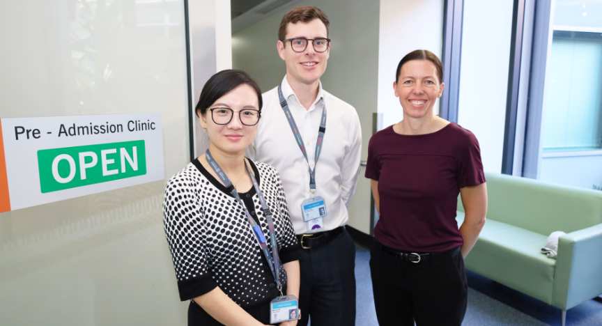 Three healthcare professionals stand in front of a sign that reads Pre-Admission Clinic OPEN in a hospital setting.