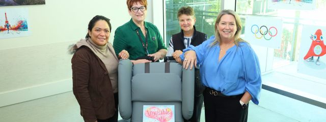 Four people stand around a grey medical device labeled Invisible Hug in a room with Olympic-themed decorations.