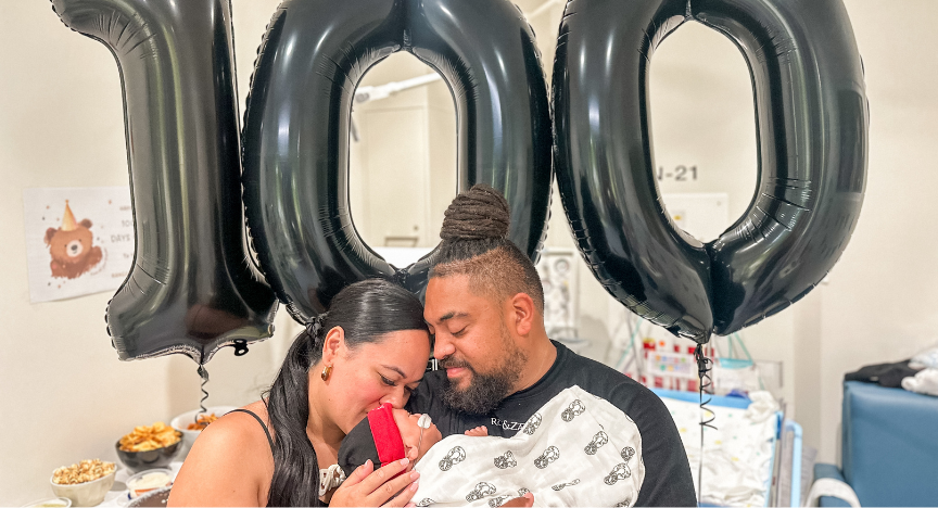 Two people holding a red object, standing in front of large black balloons forming the number 100 in a room with food and decorations.