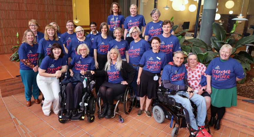 A group of people positioned in a tight group smiling at the camera in a courtyard wearing a branded t-shirt with the words Stronger Together printed on the front. The group represent hospital clinicians, hospital executive, consumers with a lived experience with spinal injuries, representatives from stakeholder Spinal Life Australia, and Mary Freer from Compassion Revolution.