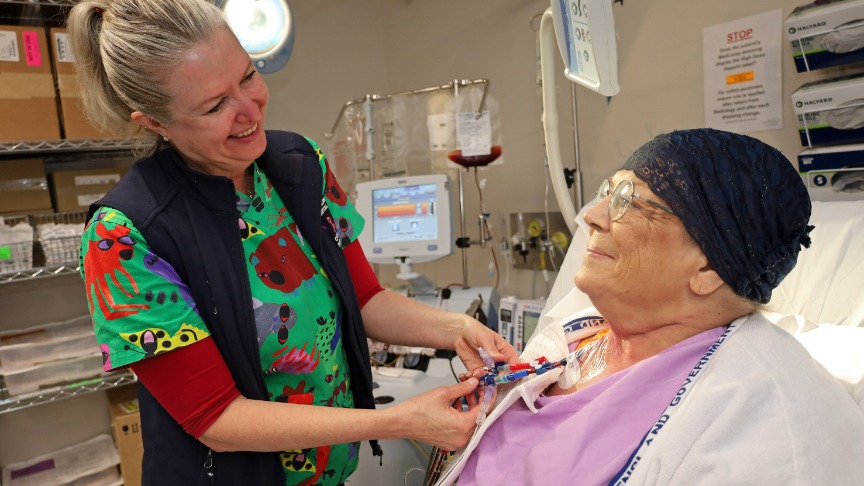 A female nurse in colourful scrubs checks the infusion lines of a female lymphoma patient sitting in bed wearing a purple smock, glasses and a head wrap in the apheresis room next to a centrifuge machine