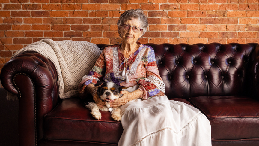 An elderly woman with short grey hair and glasses sitting on a brown leather couch and holding a small dog