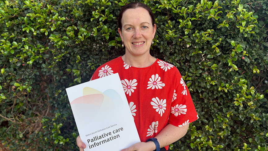 A woman wearing a red top, standing in front of a green hedge holding a document titled Palliative Care Information. She has brown hair and a smile on her face.
