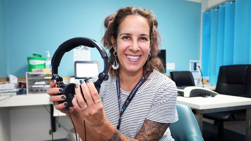 A female aboriginal clinician in a clinic room smiles at the camera holding some headphones