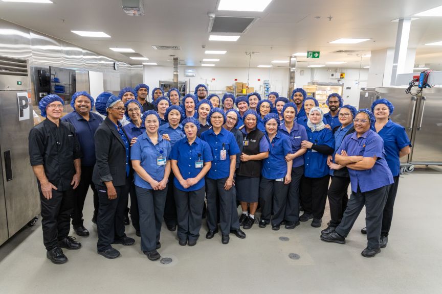 Large group of operation staff wearing blue uniform shirts and hair nets standing in a kitchen