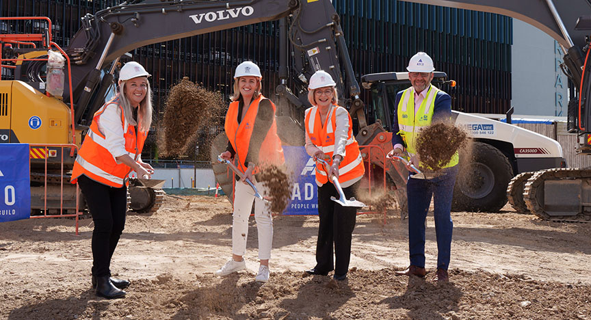 Four people in safety gear and hard hats participate in a groundbreaking ceremony with shovels, in front of construction equipment.
