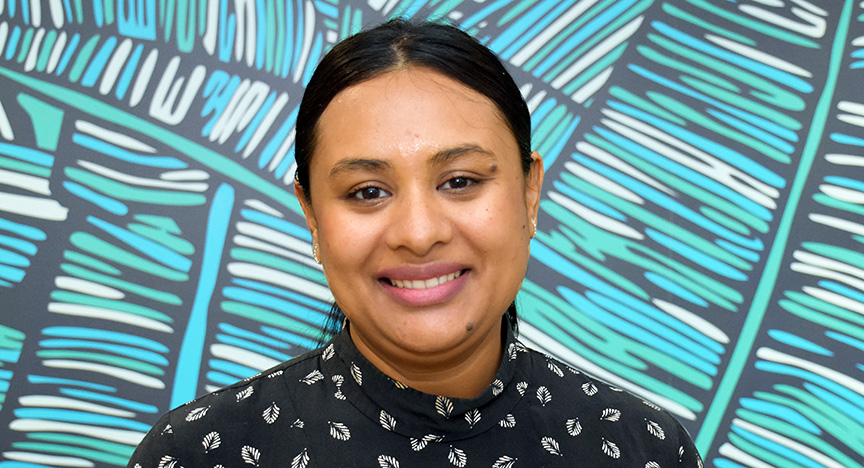 A person stands in front of a colorful, abstract background with blue and green patterns, wearing a dark shirt with a white leaf design.