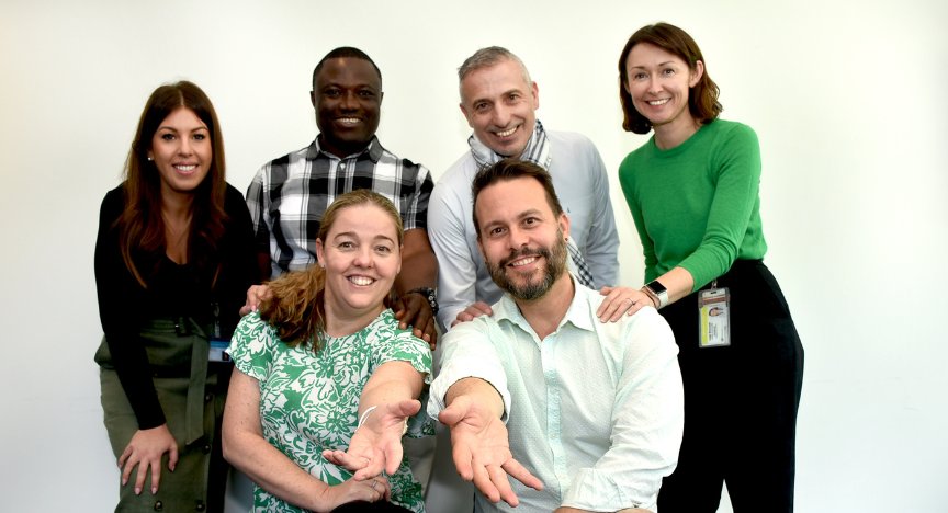 A group of six people, some seated and some standing, pose together in a friendly manner, extending their hands towards the camera.