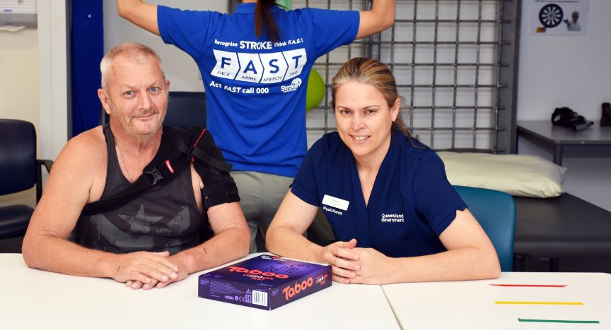 A person with an arm sling and a healthcare worker sit at a table with a Taboo game box. A third person stands behind them wearing a FAST stroke awareness shirt.