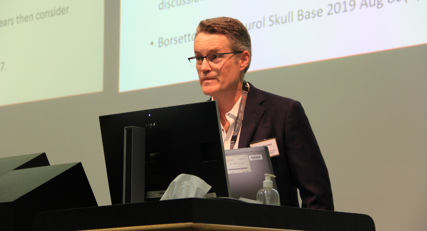 A person stands at a podium, presenting at a conference. A screen behind displays text and references.