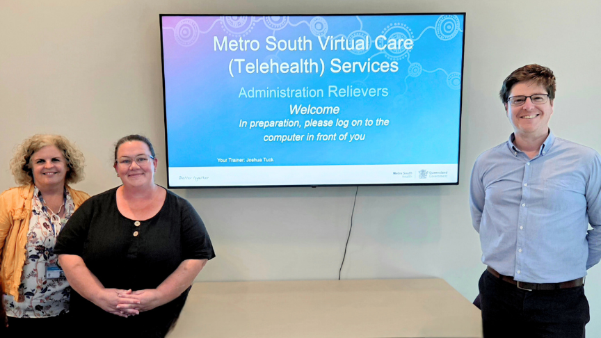 Three people stand in front of a presentation slide reading Metro South Virtual Care (Telehealth) Services Administration Relievers Welcome. In preparation, please log in to the computer in front of you.